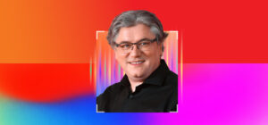 Smiling middle-aged man with gray hair and glasses in a black shirt, centered in a white-framed portrait on a vibrant red, pink, and blue gradient background.