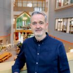 Portrait of a man in a navy shirt standing in a modern office atrium with glass meeting rooms and timber beams in the background.